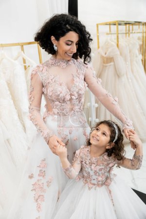delightful middle eastern bride in floral wedding dress holding hands with happy girl in cute attire in bridal salon, shopping, special moment, mother and daughter, happiness, togetherness 