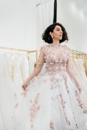 bezaubernde und nahöstliche Frau probiert wunderschöne und florale Hochzeitskleid innerhalb von luxuriösen Brautsalon, Einkaufen, Braut-zu-sein, verschwommenes weißes Kleid auf dem Hintergrund, wegschauen 