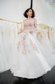 full length of brunette and middle eastern woman with wavy hair trying on gorgeous and floral wedding dress inside of luxurious bridal salon, shopping, bride-to-be,  blurred white gown Stickers #658421794