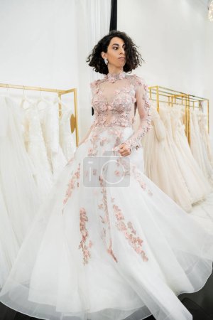 delightful and middle eastern woman with wavy hair trying on elegant and floral wedding dress inside of luxurious bridal salon, shopping, bride-to-be,  blurred white gown on background