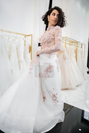 Photo for Charming and middle eastern woman with wavy hair trying on elegant and floral wedding dress with train inside of luxurious bridal salon, shopping, bride-to-be,  blurred white gown, looking away - Royalty Free Image