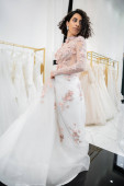 charming and middle eastern woman with wavy hair trying on elegant and floral wedding dress with train inside of luxurious bridal salon, shopping, bride-to-be,  blurred white gown, looking away   Poster #658421814