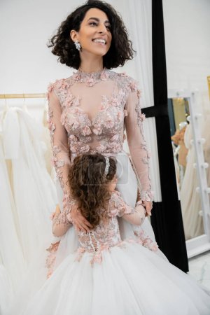 cute girl in floral attire hugging cheerful mother with brunette hair standing in wedding dress near blurred white gown inside of luxurious bridal salon, shopping, bride-to-be
