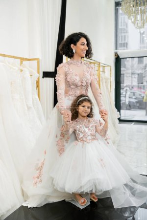 Nettes Mädchen aus dem Nahen Osten in floraler Kleidung, Händchen haltend mit einer fröhlichen Frau, die im Hochzeitskleid in der Nähe eines verschwommenen weißen Kleides im Brautsalon steht, einkaufen, zukünftige Braut, Mutter und Tochter 