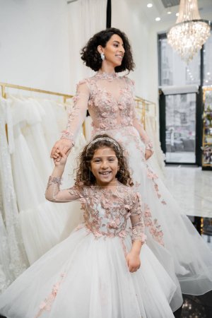 Moyen-Orient fille en tenue florale tenant la main avec femme gaie debout dans la robe de mariée près floue robe blanche à l'intérieur du salon de mariée luxueux, shopping, future mariée, mère et fille 