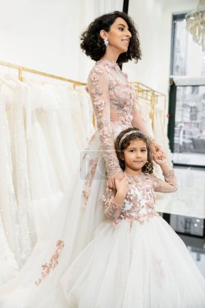 middle eastern girl in floral attire holding hands with cheerful mother standing in wedding dress near blurred white gown inside of luxurious bridal salon, shopping, bride-to-be, bonding 