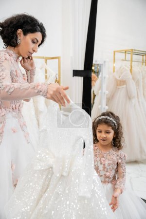 brunette middle eastern bride in floral wedding gown helping to choose dress for her little daughter in bridal salon around white tulle fabrics, process of preparation, togetherness 