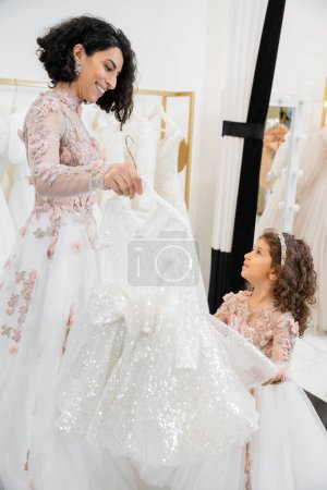 happy and brunette middle eastern bride in floral wedding gown helping to choose dress for her little daughter in bridal salon around white tulle fabrics, process of preparation, togetherness 