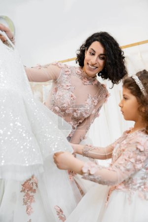 charming and brunette middle eastern woman in floral wedding gown helping to choose dress for her little daughter in bridal salon around white tulle fabrics, process of preparation, togetherness 