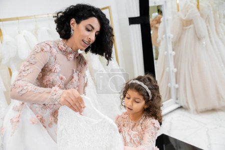 Téléchargez les photos : Brunette mariée du Moyen-Orient en robe de mariée florale aidant à choisir la robe pour sa jolie petite fille dans la boutique nuptiale autour de tissus en tulle blanc, processus de préparation, ensemble - en image libre de droit