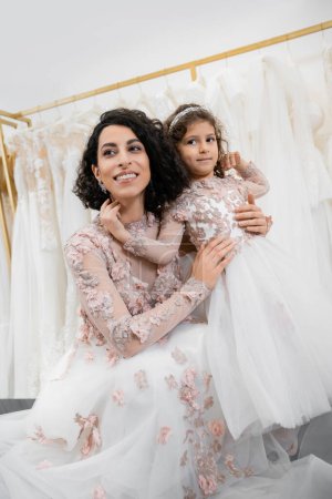 besonderer Moment, entzückende mittelöstliche Braut im floralen Brautkleid, die ihre kleine Tochter im Brautsalon um weiße Tüll-Stoffe umarmt, Brautshopping, Zweisamkeit 
