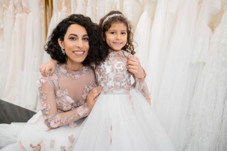 moment spécial, heureuse femme du Moyen-Orient en robe de mariée florale assise et embrassant sa petite fille dans un salon nuptial autour de tissus en tulle blanc, shopping nuptial, convivialité, future mariée 
