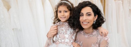 special moment, cheerful middle eastern bride in floral wedding gown sitting and embracing her little daughter in bridal salon around white tulle fabrics, bridal shopping, togetherness, banner 