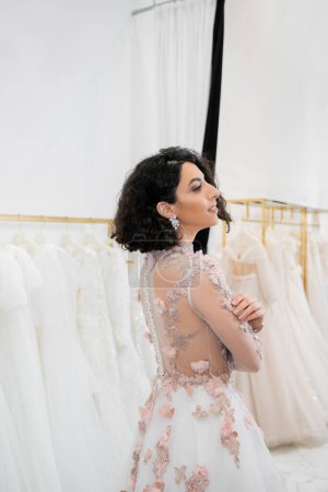 stunning middle eastern and brunette woman with wavy hair standing in gorgeous and floral wedding dress inside of luxurious bridal salon around white tulle fabrics, bridal shopping, looking away 