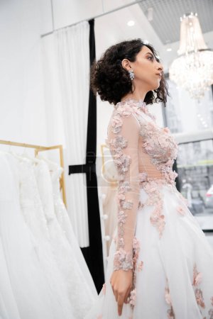 bride-to-be, stunning middle eastern and brunette woman with wavy hair standing in gorgeous and floral wedding dress inside of luxurious bridal salon around white tulle fabrics, bridal shopping 