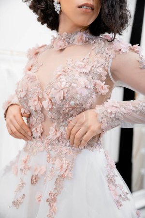 Ausgeschnittene Ansicht einer brünetten Frau, die in einem wunderschönen und floralen Hochzeitskleid in einem luxuriösen Salon um weiße Tüll-Stoffe steht, Brautshopping, zukünftige Braut, Details 