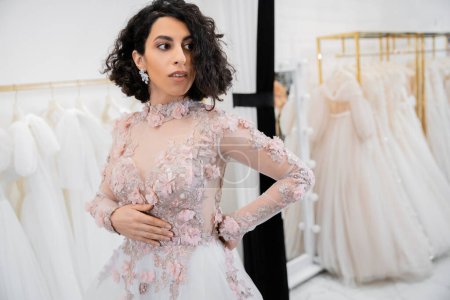 bride-to-be, alluring middle eastern and brunette woman with wavy hair standing in gorgeous and floral wedding dress inside of luxurious salon around white tulle fabrics, bridal shopping 