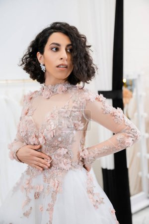 bride-to-be, brunette middle eastern woman with wavy hair standing in gorgeous and floral wedding dress inside of luxurious salon around white tulle fabrics, bridal shopping, sophisticated 