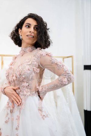 cheerful middle eastern and brunette woman with wavy hair standing in elegant and floral wedding dress and looking away inside of luxurious salon around white tulle fabrics, bridal shopping 