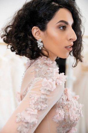 retrato de la encantadora mujer de Oriente Medio con el pelo ondulado de pie en vestido de novia hermoso y floral y mirando hacia el interior del lujoso salón alrededor de telas de tul blanco, compras nupciales 