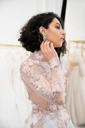delightful middle eastern woman with wavy hair standing in gorgeous and floral wedding dress and touching earring inside of luxurious salon around white tulle fabrics, bridal shopping 