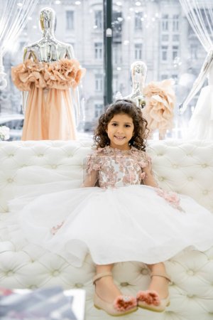 Photo for Cheerful middle eastern girl with curly hair sitting in floral dress on white couch inside of luxurious wedding salon, smiling kid, tulle skirt, bridal, blurred mannequin on background - Royalty Free Image