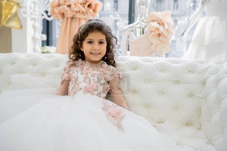 alegre chica de Oriente Medio con el pelo rizado sentado en vestido floral en el sofá blanco y sonriendo dentro de lujoso salón de bodas, niño sonriente, falda de tul, fondo borroso 