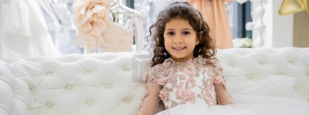 happy middle eastern girl with curly hair sitting in floral dress on white couch and smiling inside of luxurious wedding salon, smiling kid, tulle skirt, blurred background, banner 