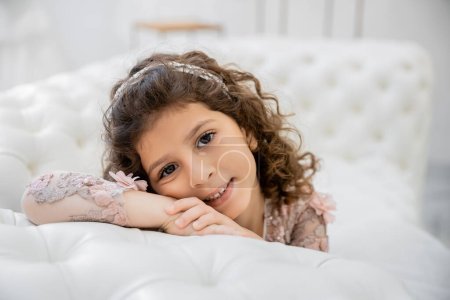 Porträt eines positiven mittelöstlichen Mädchens mit brünetten lockigen Haaren, das in einem floralen Kleid posiert und auf einer weißen Couch in einem luxuriösen Hochzeitssalon lehnt, lächelndes Kind, verschwommener Hintergrund 