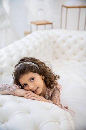 portrait of cheerful middle eastern girl with brunette curly hair posing in floral dress and leaning on white couch inside of luxurious wedding salon, smiling kid, blurred background, joy 