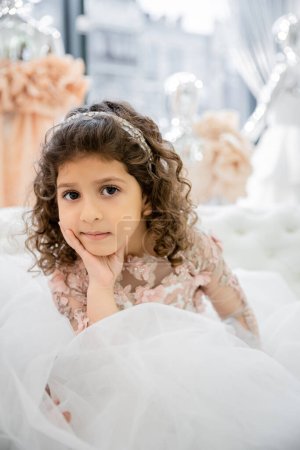 Porträt des netten kleinen Mädchens aus dem Nahen Osten mit lockigem Haar, das in einem floralen Kleid auf einer weißen Couch in einem luxuriösen Hochzeitssalon sitzt, Tüllrock, verschwommener Hintergrund, Blick in die Kamera 