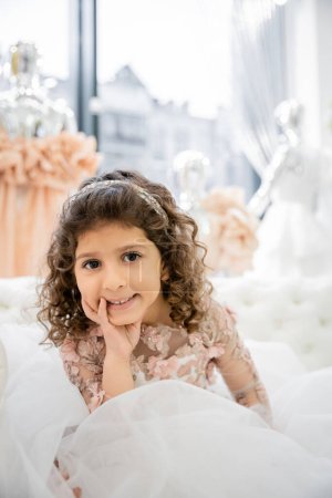 cheerful middle eastern girl with curly hair posing in floral dress with tulle skirt and sitting on white couch inside of luxurious wedding salon, smiling kid, blurred background 