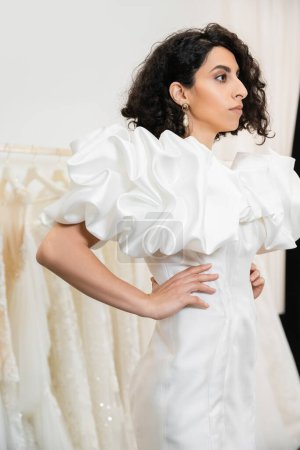 brunette middle eastern bride with brunette and wavy hair posing with hands on hips in trendy wedding dress with puff sleeves and ruffles in bridal salon next to tulle fabrics