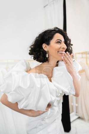 Photo for Radiant middle eastern bride with brunette wavy hair laughing, covering mouth and posing in trendy wedding dress with puff sleeves and ruffles in bridal salon, fashion - Royalty Free Image
