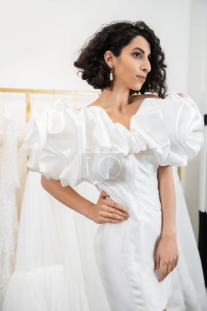 brunette middle eastern bride with brunette and wavy hair posing with hand on hip in stylish wedding dress with puff sleeves and ruffles in bridal boutique next to tulle fabrics, delightful woman 