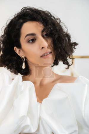 portrait of brunette middle eastern bride with wavy hair posing in trendy wedding dress with puff sleeves and ruffles in bridal boutique and looking away, elegant woman 