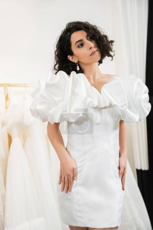 elegante mittelöstliche Braut mit brünetten und welligen Haaren posiert im trendigen Hochzeitskleid mit Puffärmeln und Rüschen in der Brautboutique neben Tüll-Stoffen, elegante Frau 