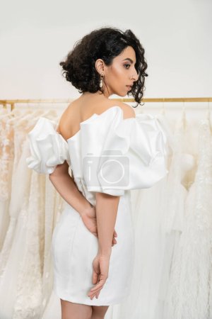 stunning middle eastern bride with brunette and wavy hair posing in stylish wedding dress with puff sleeves and ruffles in bridal boutique next to tulle fabrics, elegant woman  magic mug #658422770