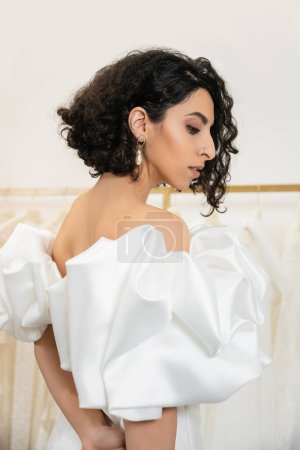 Photo for Brunette middle eastern woman with brunette and wavy hair posing in trendy wedding dress with puff sleeves and ruffles in bridal boutique and looking away, elegant bride, shopping - Royalty Free Image