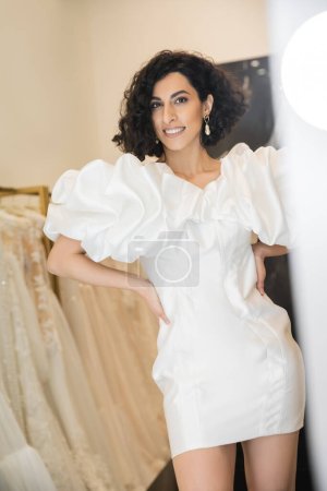 glückliche Frau aus dem Nahen Osten mit brünetten welligen Haaren beim Anprobieren eines Brautkleides mit Puffärmeln und Rüschen in der Nähe eines Spiegels in einer Brautboutique neben Tüll-Stoffen, Reflexion, Shopping, Hände an den Hüften