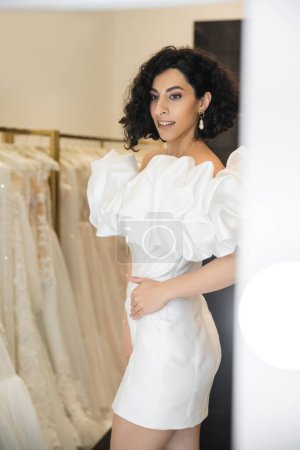 smiling middle eastern woman with brunette and wavy hair trying on trendy wedding dress with puff sleeves and ruffles near mirror in bridal boutique next to tulle fabrics, reflection, shopping 
