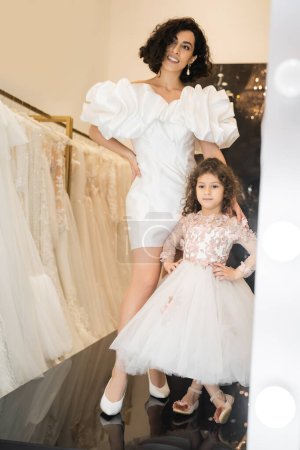 joyful middle eastern bride with wavy hair posing in trendy wedding dress with puff sleeves and ruffles near cute little daughter in floral attire and white bridal gown in boutique  