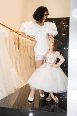 happy brunette middle eastern woman with wavy hair posing in trendy wedding dress with puff sleeves and ruffles near cute little daughter in floral attire and white bridal gown in boutique  