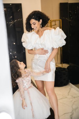 happy and cute middle eastern little girl in floral attire looking at bride with wavy hair standing in trendy wedding dress with puff sleeves and ruffles in bridal boutique, mother and daughter 