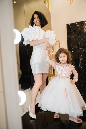 happy middle eastern bride with wavy hair standing in trendy wedding dress with puff sleeves and ruffles while looking at mirror near cute little daughter in floral attire in bridal boutique  