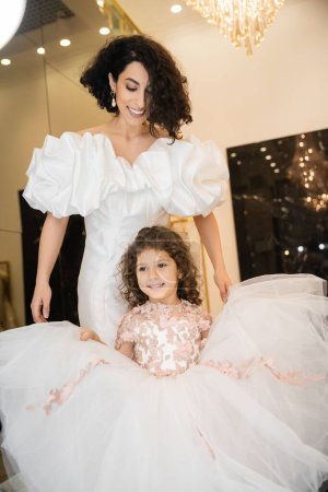 joyful middle eastern little girl in floral attire holding tulle skirt and looking at mirror near bride with wavy hair standing in wedding dress with puff sleeves and ruffles in bridal boutique 