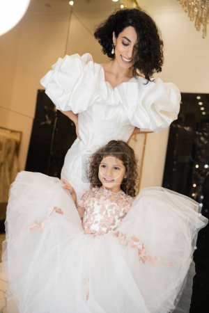 happy little girl in floral attire holding tulle skirt and looking away near  middle eastern mother with wavy hair standing in white wedding dress with puff sleeves and ruffles in bridal boutique 