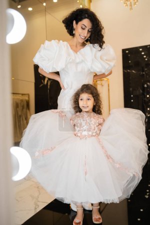 happy little girl in floral dress holding tulle skirt and looking at mirror near  middle eastern mother with wavy hair standing in white wedding gown with puff sleeves and ruffles in bridal boutique