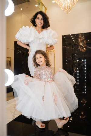 happy girl in dress holding tulle skirt and looking at mirror near charming middle eastern mother with wavy hair standing in white wedding gown with puff sleeves and ruffles in bridal boutique