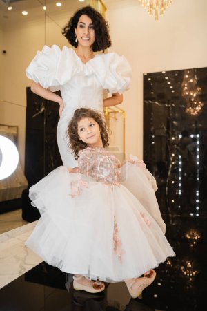 Charmante Braut aus dem Mittleren Osten mit brünetten Haaren, die in einem weißen Brautkleid mit Puffärmeln und Rüschen steht und in den Spiegel neben der Tochter blickt, die einen Tüllrock im Brautladen hält 